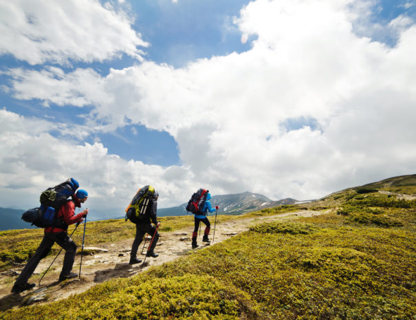 A group of people with backpacks walking along the road. The road strewns with stones. There are mountains on the horizon. The sky is blue and cloudy.